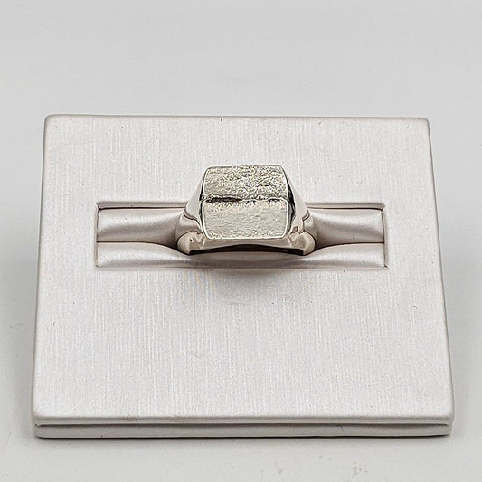 A silver-toned ring with a textured, block-like top and smooth band, displayed on a brushed silver rectangular stand against a light gray background.