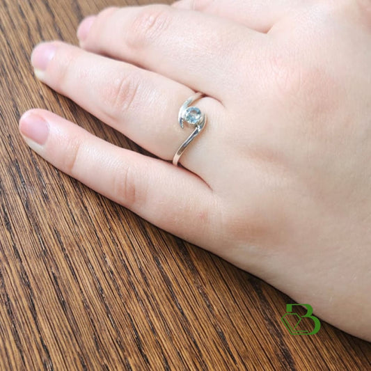 Sterling Silver Bypass ring set with a faceted Santa Maria Aquamarine gemstone on the ring finger f a woman's left hand against a wooden background.
