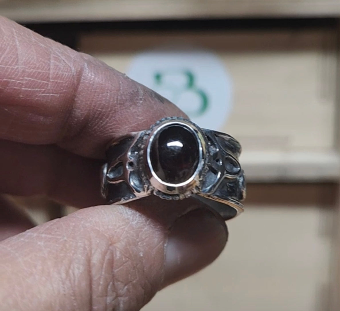A hand holding a silver-toned ring with a large oval dark reddish-brown stone, photographed against a blurred indoor background.