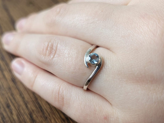 Sterling Silver Bypass ring set with a faceted Santa Maria Aquamarine gemstone on the ring finger of a woman's left hand against a blurred  background.