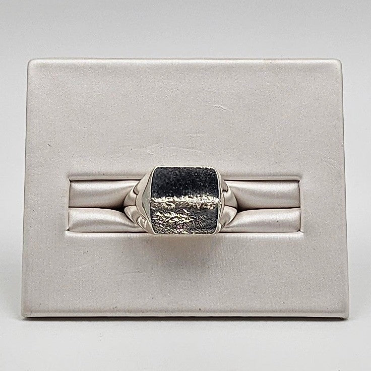 A silver ring with a rectangular, textured dark stone, displayed on a white padded jewelry stand.