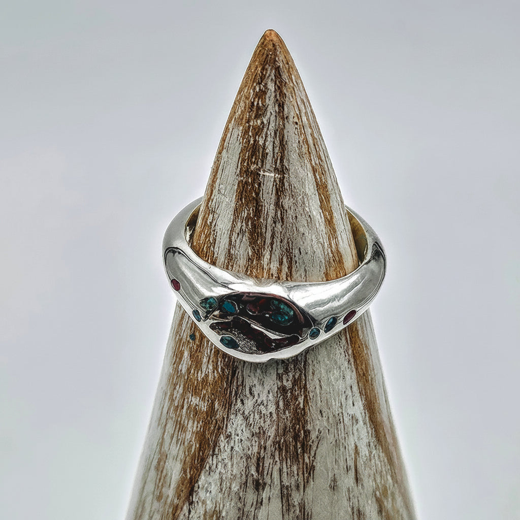 A sterling silver band ring with a polished organic-shaped setting featuring small dark and blue-green stones, displayed on a textured wooden cone stand against a light gray background.