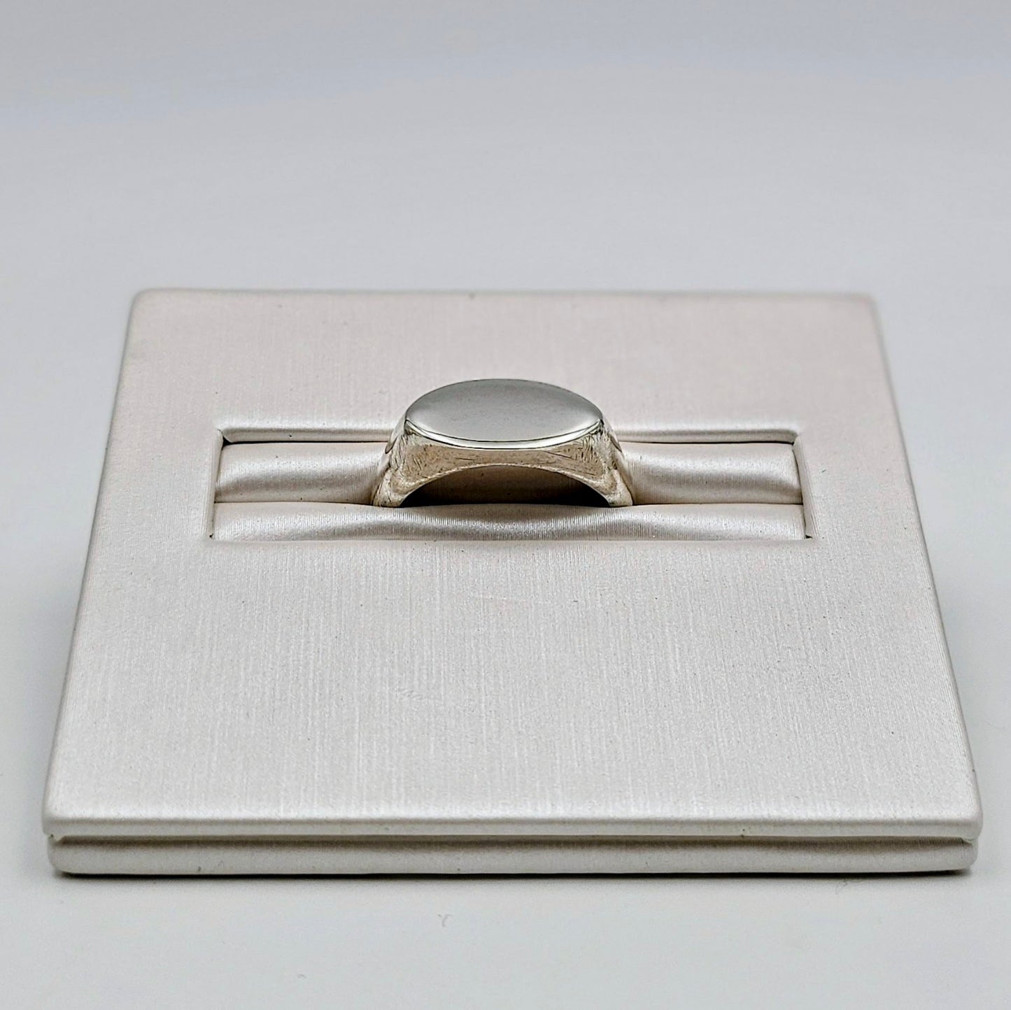 A silver signet ring displayed in a textured white presentation box on a light gray background.