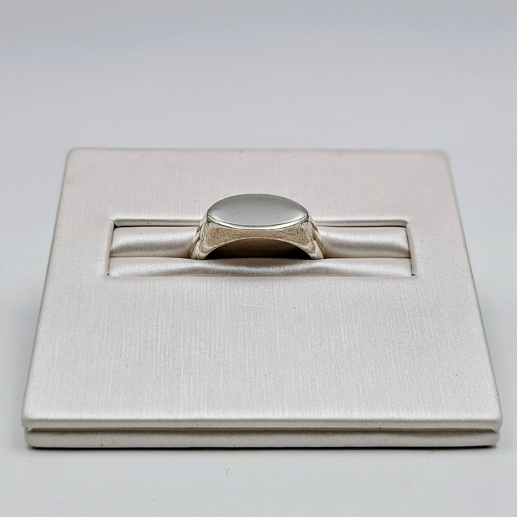 A silver signet ring displayed in a textured white presentation box on a light gray background.
