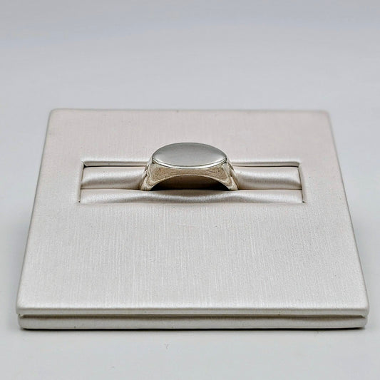 A silver signet ring displayed in a textured white presentation box on a light gray background.