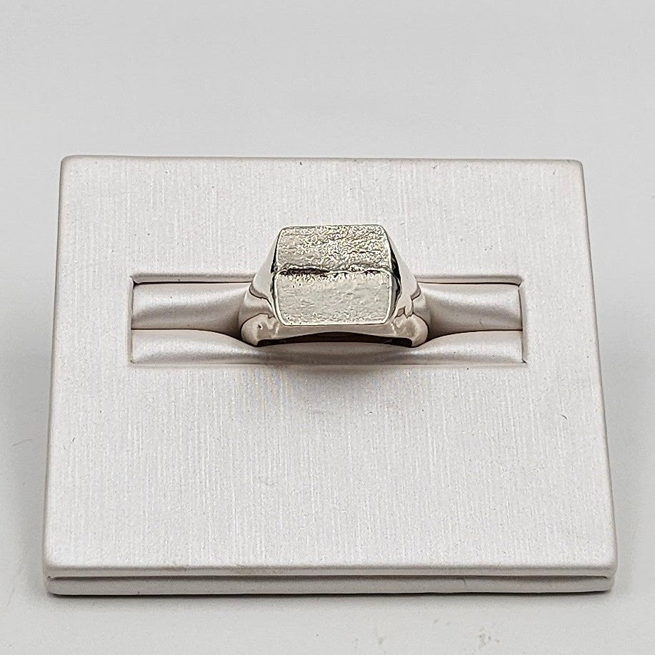 A silver-toned ring with a textured, block-like top and smooth band, displayed on a brushed silver rectangular stand against a light gray background.