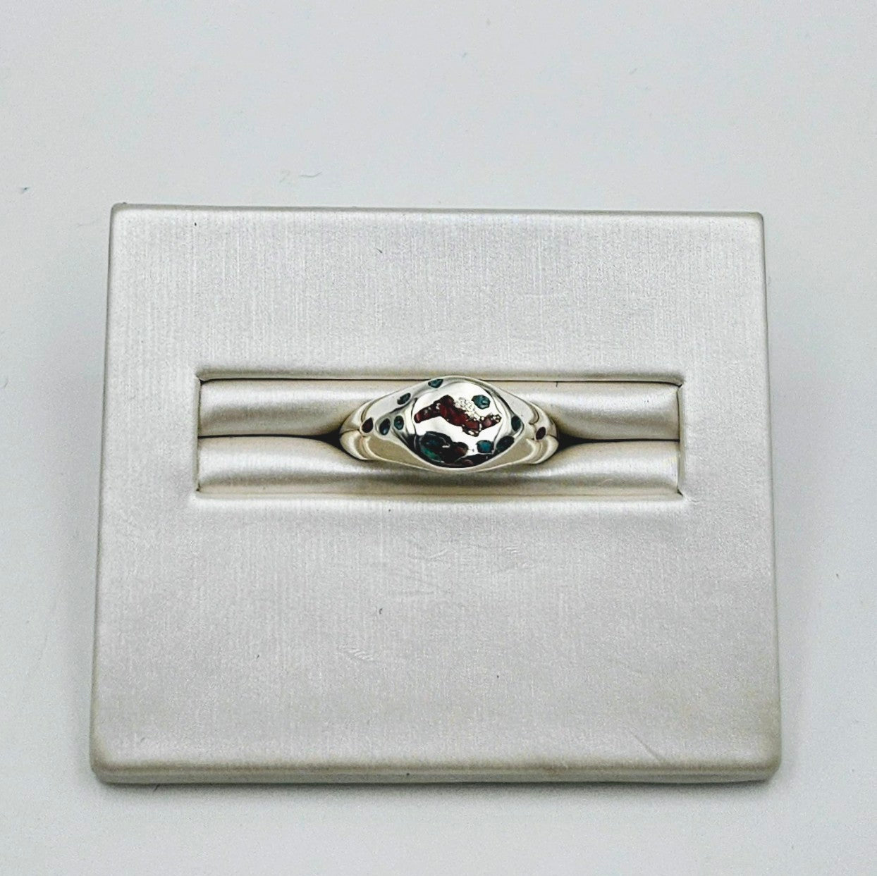 A silver-toned ring displayed in a rectangular metal jewelry box on a plain light background.