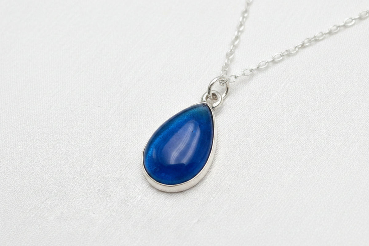 A silver teardrop pendant necklace with a vibrant blue marbled gemstone, displayed on a textured white fabric background.