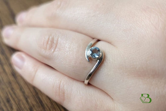 Sterling Silver Bypass ring set with a faceted Santa Maria Aquamarine gemstone on the ring finger of a woman's left hand against a blurred  background.